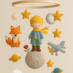 May include: A felt baby mobile with a yellow-haired prince standing on a gray moon, a fox, a plane, and stars. The prince is wearing a blue jacket, a yellow scarf, and green pants. The mobile is suspended from a wooden ring.