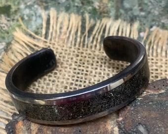 May include: A dark gray cuff bracelet with a textured, black center band and a silver-colored border. The bracelet is open-ended and rests on a rough, textured surface. The jewelry is displayed on a burlap cloth.