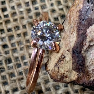 Puede incluir: Un anillo de oro rosa con una gema redonda, clara y facetada, sujeta por un engaste de cuatro garras. La banda tiene un acabado martillado y texturizado, y está colocada junto a una roca rugosa, porosa, de color marrón y bronceado.
