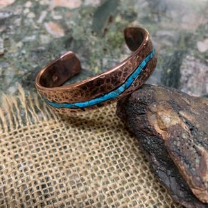 May include: A copper cuff bracelet featuring a hammered, textured surface and a central horizontal inlay of bright blue turquoise stone. The metal has a warm, reddish-brown metallic finish with an open back design for easy adjustment on the wrist.