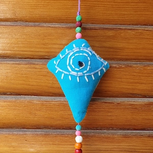 May include: A turquoise fabric eye charm with white stitched details and a beaded tassel. The charm is diamond-shaped with a downward-pointing triangle. The tassel includes wooden beads and colorful accents, suspended from a purple string.