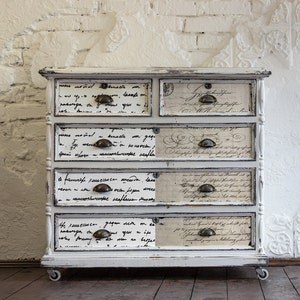 May include: White painted wooden dresser with six drawers. Each drawer front is decorated with a different handwritten text pattern. The dresser has brass handles and sits on four small wheels.