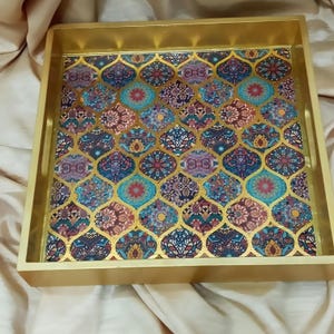 May include: A gold-colored tray with a decorative pattern of colorful geometric shapes. The tray has a raised edge and a smooth surface.