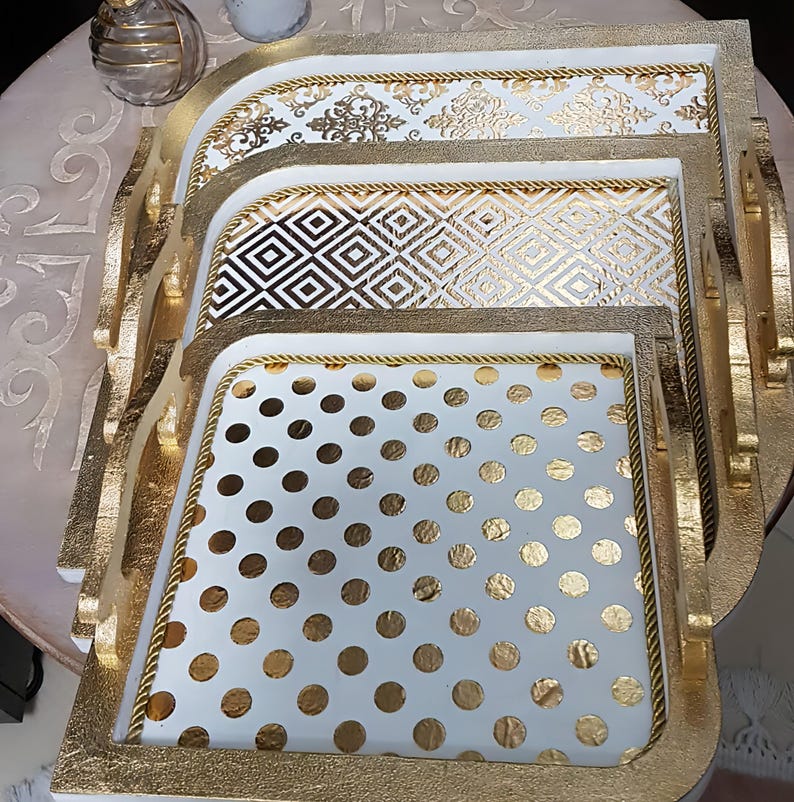 May include: Set of three white wooden trays with gold trim and different patterns. The top tray has a damask pattern, the middle tray has a geometric pattern, and the bottom tray has a polka dot pattern.