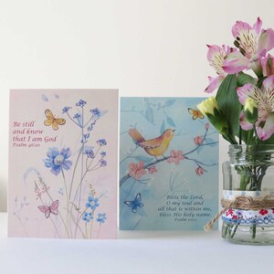 Könnte beinhalten: Zwei Grußkarten mit Aquarell-Illustrationen und inspirierenden Zitaten aus der Bibel. Die erste Karte zeigt einen rosa Hintergrund mit blauen Blumen und Schmetterlingen und den Text "Sei still und wisse, dass ich Gott bin, Psalm 46:10". Die zweite Karte zeigt einen blauen Hintergrund mit einem gelben Vogel, der auf einem Zweig mit rosa Blüten sitzt, und den Text "Lobe den Herrn, meine Seele, und alles, was in mir ist, lobe seinen heiligen Namen. Psalm 103:1".