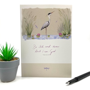 Könnte beinhalten: Eine beige Karte mit einer Aquarell-Illustration eines grauen Reihers, der in einem Teich mit rosa Seerosen und Libellen steht. Der Text "Be still, and know that I am God" ist in lila Kursivschrift mit "Psalm 46:10" darunter geschrieben.