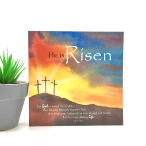 May include: Square art print featuring a watercolour sunset scene with three crosses on a hill. The text "He is Risen" is at the top, with a Bible verse below. A small potted plant is on the left.