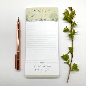 May include: A green and white notepad with a lined interior. The notepad features a nature scene with a heron, dragonflies, and water lilies. The bottom of the notepad has the text "Be still and know that I am God" and "Psalm 46:10".