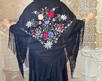 May include: A black triangular shawl featuring vibrant floral embroidery in red, blue, yellow, and white. The shawl is adorned with long, black fringe along the edges, creating a textured appearance. The fabric is smooth and dark, highlighting the colorful thread work.
