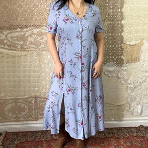 Op de afbeelding: Lichtblauwe jurk met knopen en een bloemenpatroon van roze en witte bloemen. De jurk heeft korte mouwen en een split aan de voorkant. De persoon draagt bruine cowboylaarzen.