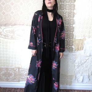 May include: A long, black robe with a floral pattern in pink, purple, and yellow. The robe has pink trim and is worn over a black dress. The background features a patterned wall and a red rug.