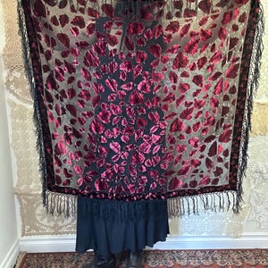 May include: A sheer black shawl with a pattern of burgundy velvet leaves. The shawl has a black fringe border. The leaves are scattered across the shawl, creating a striking contrast against the sheer background. The shawl is a fashion accessory.