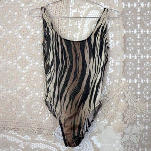 May include: A vintage swimsuit with a zebra print in black, tan, and brown. The one-piece has a scoop neckline and is suspended on a metal hanger. The backdrop is a cream-coloured lace.