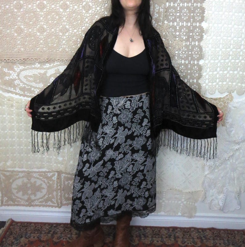 May include: A black shawl with a velvet texture and fringe trim. The shawl has a geometric pattern with red and purple accents. The model is wearing a black top and a black and gray floral skirt.