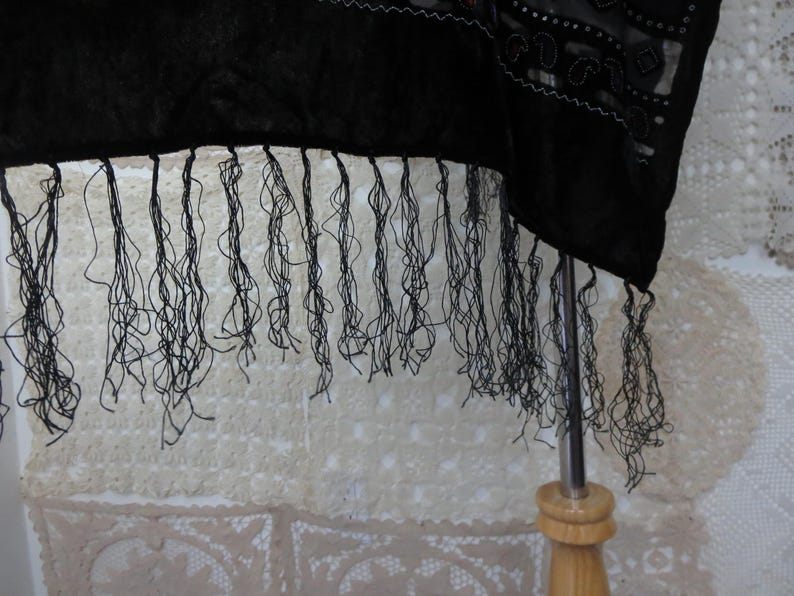 May include: A black shawl with long, delicate fringe. The fabric appears to be a dark, possibly velvet material, with intricate detailing along the edge. The fringe hangs down, creating a textured effect.