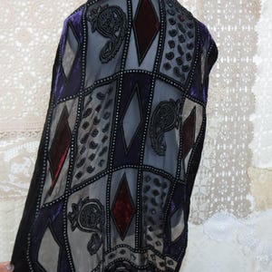 May include: A decorative shawl with a geometric pattern of squares and diamonds in shades of purple, gray, and burgundy. The shawl features intricate paisley and dot designs, with a black fringe along the bottom edge.