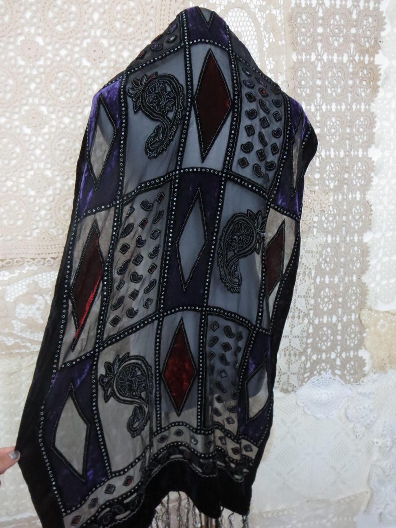 May include: A decorative shawl with a geometric pattern of squares and diamonds in shades of purple, gray, and burgundy. The shawl features intricate paisley and dot designs, with a black fringe along the bottom edge.