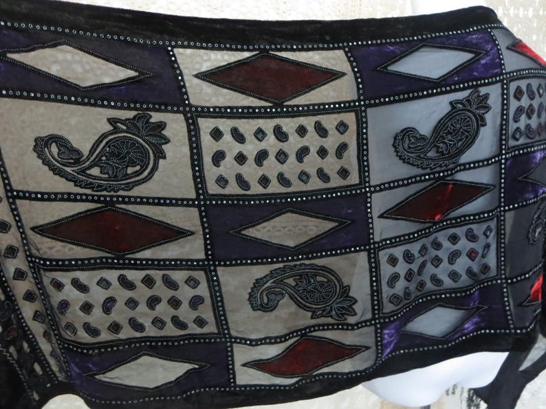 May include: A black, sheer scarf with a patchwork design. The scarf features squares in gray, purple, and burgundy, adorned with paisley and diamond patterns. The edges are trimmed with small, silver beads.