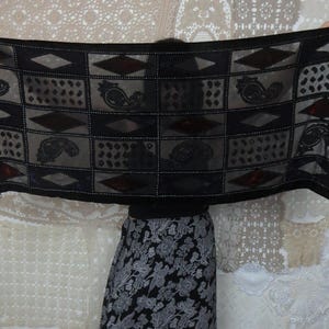 May include: A black scarf with a geometric pattern of squares, diamonds, and paisley designs. The scarf has a sheer, lightweight fabric with a fringe trim at the ends. The design includes dark red accents.
