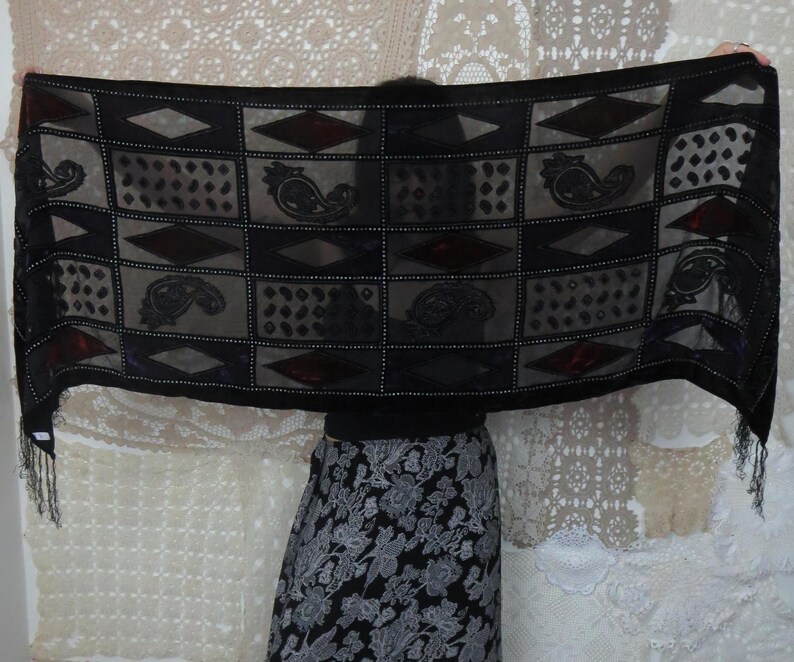 May include: A black scarf with a geometric pattern of squares, diamonds, and paisley designs. The scarf has a sheer, lightweight fabric with a fringe trim at the ends. The design includes dark red accents.
