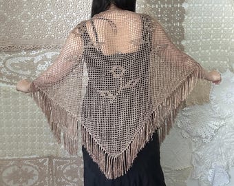 May include: A tan crocheted shawl with a fringe trim. The shawl is worn over a black dress. The shawl has a decorative pattern on the back. The background features various crocheted items.