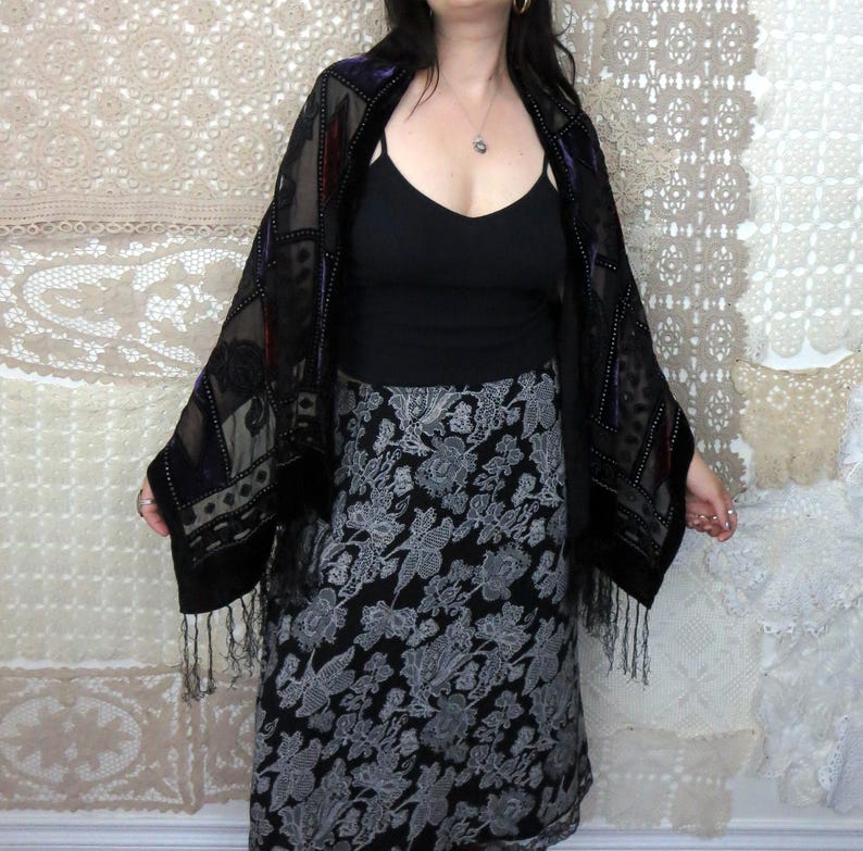 May include: Black sheer shawl with a geometric pattern and fringe. The shawl is worn over a black top and a black and white floral skirt. The shawl has a velvet trim and is embellished with beads.