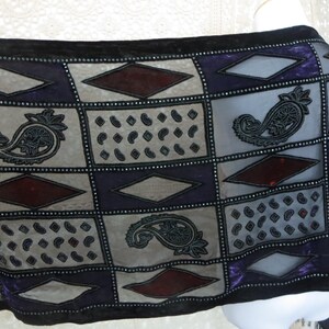 May include: A sheer scarf with a patchwork design featuring paisley, diamond, and dot patterns in gray, purple, and burgundy. The scarf is trimmed with black velvet and embellished with small, silver-colored beads.