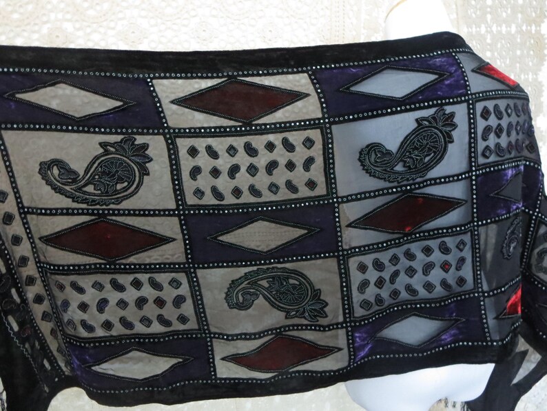 May include: A sheer scarf with a patchwork design featuring paisley, diamond, and dot patterns in gray, purple, and burgundy. The scarf is trimmed with black velvet and embellished with small, silver-colored beads.