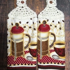 Könnte beinhalten: Zwei weiße Häkel-Geschirrtücher mit braunen Knöpfen. Jedes Handtuch zeigt ein gedrucktes Design einer Kaffeekanne, einer Kaffeetasse und Kaffeebohnen auf weißem Hintergrund mit roten, braunen und goldenen Akzenten.