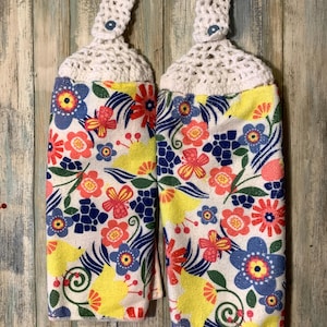 Może przedstawiać: Dwa białe ręczniki kuchenne z kwiatowym nadrukiem w odcieniach żółtego, niebieskiego, czerwonego i zielonego. Ręczniki mają na górze haftowane pętle z guzikiem.