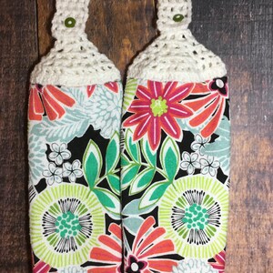 May include: Two kitchen towels with a floral print in shades of pink, green, and white on a black background. The towels have white crocheted loops at the top.