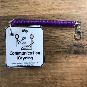 Puede incluir: Una tarjeta blanca con texto negro y una ilustraci&oacute;n simple de figura de palo. La tarjeta dice "My Communication Keyring" e incluye el nombre de la empresa y el sitio web. La tarjeta est&aacute; unida a un llavero de espiral morado.