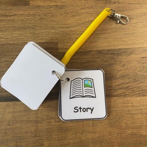 Puede incluir: Una tarjeta blanca con una ilustraci&oacute;n en blanco y negro de un libro abierto con un peque&ntilde;o sol y un campo verde en su interior. La tarjeta tiene la palabra "Story" impresa en negro debajo de la ilustraci&oacute;n. La tarjeta est&aacute; unida a un llavero enrollado amarillo.