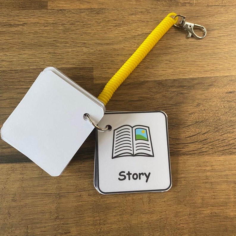 Puede incluir: Una tarjeta blanca con una ilustraci&oacute;n en blanco y negro de un libro abierto con un peque&ntilde;o sol y un campo verde en su interior. La tarjeta tiene la palabra "Story" impresa en negro debajo de la ilustraci&oacute;n. La tarjeta est&aacute; unida a un llavero enrollado amarillo.
