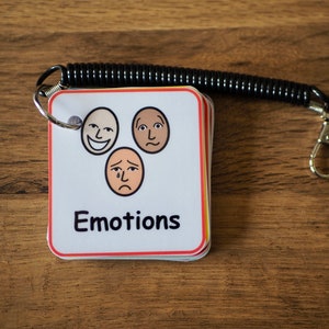 May include: A set of small, white cards with colourful borders, featuring different facial expressions. The cards are attached to a black spring coil with a silver key ring. The cards are labelled "Emotions".