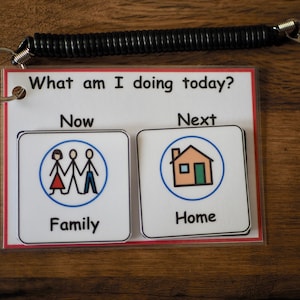 May include: A white visual schedule with a black lanyard. The schedule has two sections labelled "Now" and "Next". The "Now" section shows a family of three stick figures in a blue circle. The "Next" section shows a house in a blue circle. The text "What am I doing today?" is at the top of the schedule.