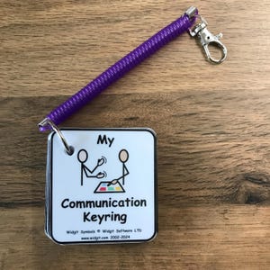 May include: A white card with black text and a black and white illustration of two stick figures communicating. The card says "My Communication Keyring" and "Widget Symbols © Widget Software LTD www.widgit.com 2002-2024". The card is attached to a purple coiled keychain with a silver metal ring and a silver metal hook.