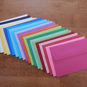 May include: A selection of colourful envelopes in a range of shades, including black, yellow, blue, green, and pink, arranged on a wooden surface. The envelopes are neatly stacked, displaying a spectrum of colours.