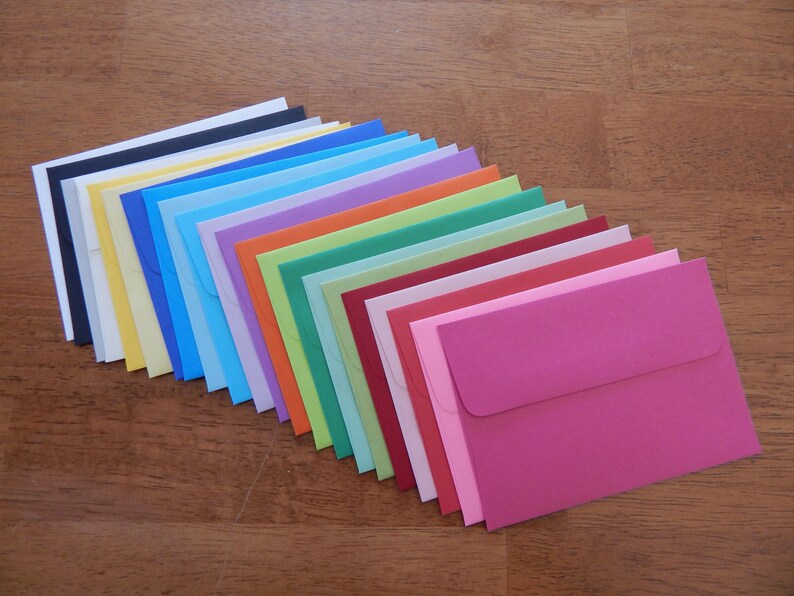 May include: A selection of colourful envelopes in a range of shades, including black, yellow, blue, green, and pink, arranged on a wooden surface. The envelopes are neatly stacked, displaying a spectrum of colours.