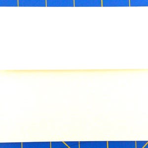 May include: A folded white paper, possibly a card or envelope, is centered on a blue cutting mat with yellow grid lines. The paper is folded horizontally, creating two distinct sections. The lighting is bright and even.