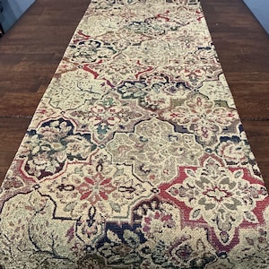 May include: A rectangular table runner featuring a repeating quatrefoil pattern in shades of deep red, navy blue, olive green, and beige. The fabric has a textured, woven appearance with intricate floral and geometric motifs throughout the design.