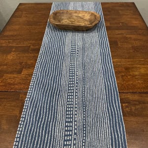 May include: A blue and white patterned table runner with a fringed edge. A wooden bowl sits atop the runner, which is placed on a dark wood table. The runner's design features vertical lines and a central grid pattern.