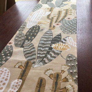 May include: A beige table runner featuring a repeating pattern of stylized cacti in shades of green, brown, and white. The runner is rectangular and appears to be made of fabric. The design includes various cactus shapes and floral elements.