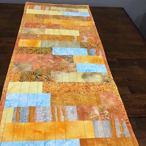May include: A rectangular table runner with a patchwork design. The runner features a mix of orange, yellow, and blue fabric blocks. The fabrics have various patterns, including floral and speckled designs. The edges are finished with a matching orange border.