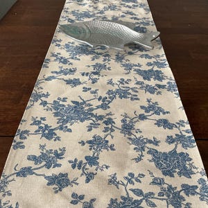 May include: A decorative table runner with a blue floral pattern on a cream background. A silver fish-shaped serving dish rests on the runner. The runner is made of fabric and is rectangular in shape. The floral design includes roses and other flowers.