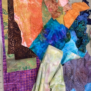 May include: A collection of assorted fabric scraps featuring various patterns and colors, including orange, blue, purple, green, and brown. The pieces display diverse textures and prints, such as marbled, grid, and organic shapes, arranged on a cutting mat.