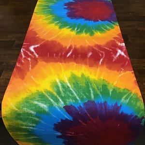 May include: A colorful tie-dye table runner. The runner features a spiral design with a vibrant array of colors, including red, orange, yellow, green, blue, and purple. The fabric is likely cotton or a similar material, suitable for home decor.