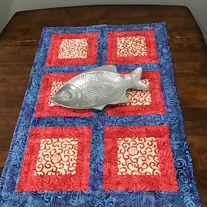 May include: A blue and red quilted table runner with a white floral pattern. A silver fish-shaped serving dish is placed on top of the runner.
