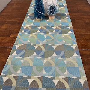 May include: A rectangular table runner with a geometric pattern in shades of blue, green, and gray. Three small decorative bottle brush Christmas trees sit on top. The runner is made of a woven fabric.