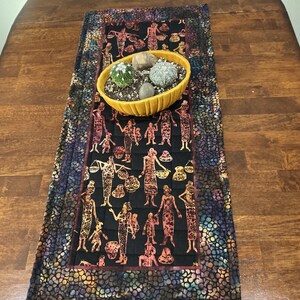 May include: A rectangular fabric table runner featuring a central black panel with a repeating orange and brown pattern of figures and pottery, bordered by a dark multi-colored mosaic print. A yellow oval bowl with cacti sits on top.
